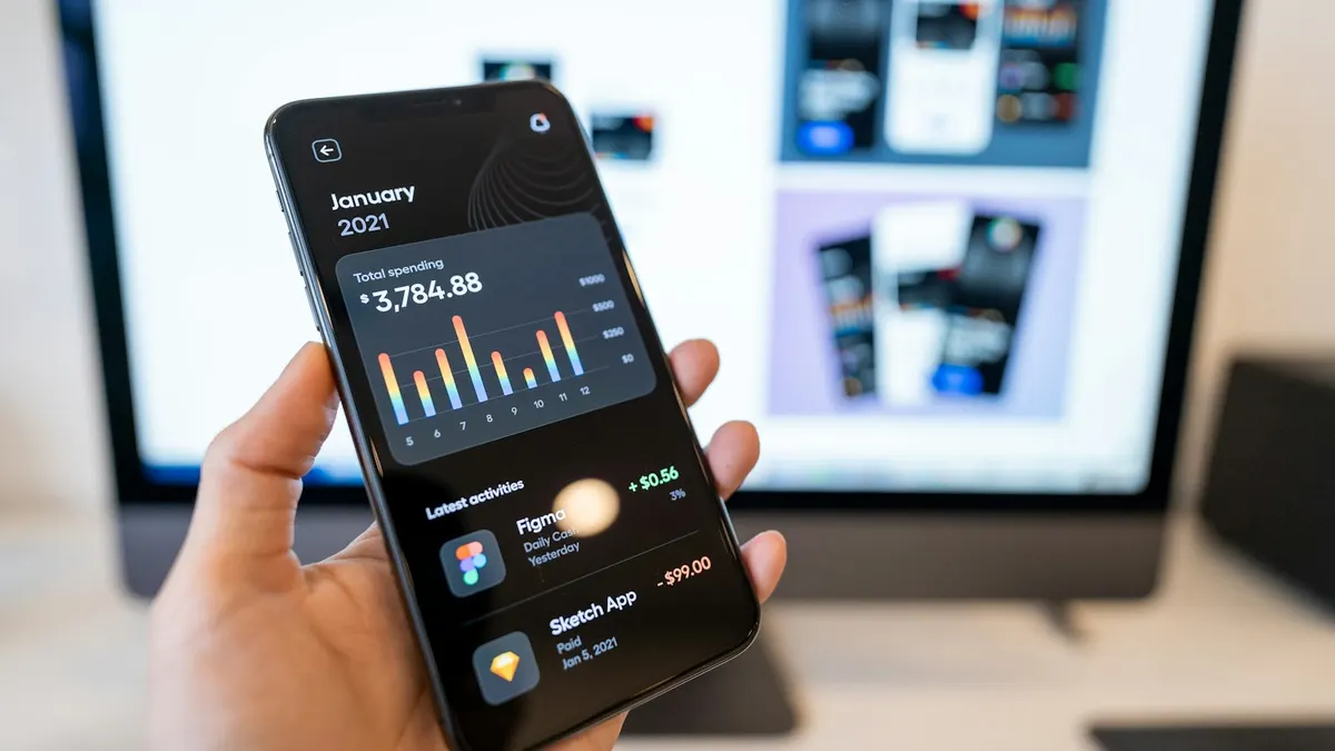 A hand holding a phone with a finished app next to a monitor showing design screens