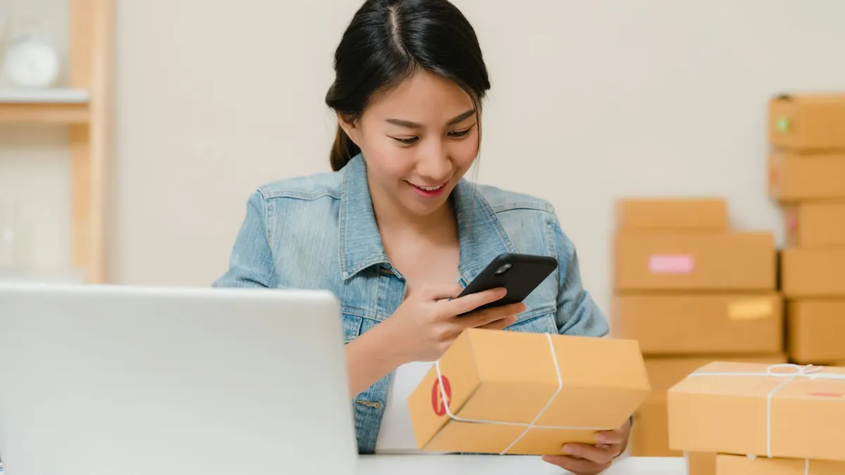 Woman checking a package with a phone beside a laptop and boxes